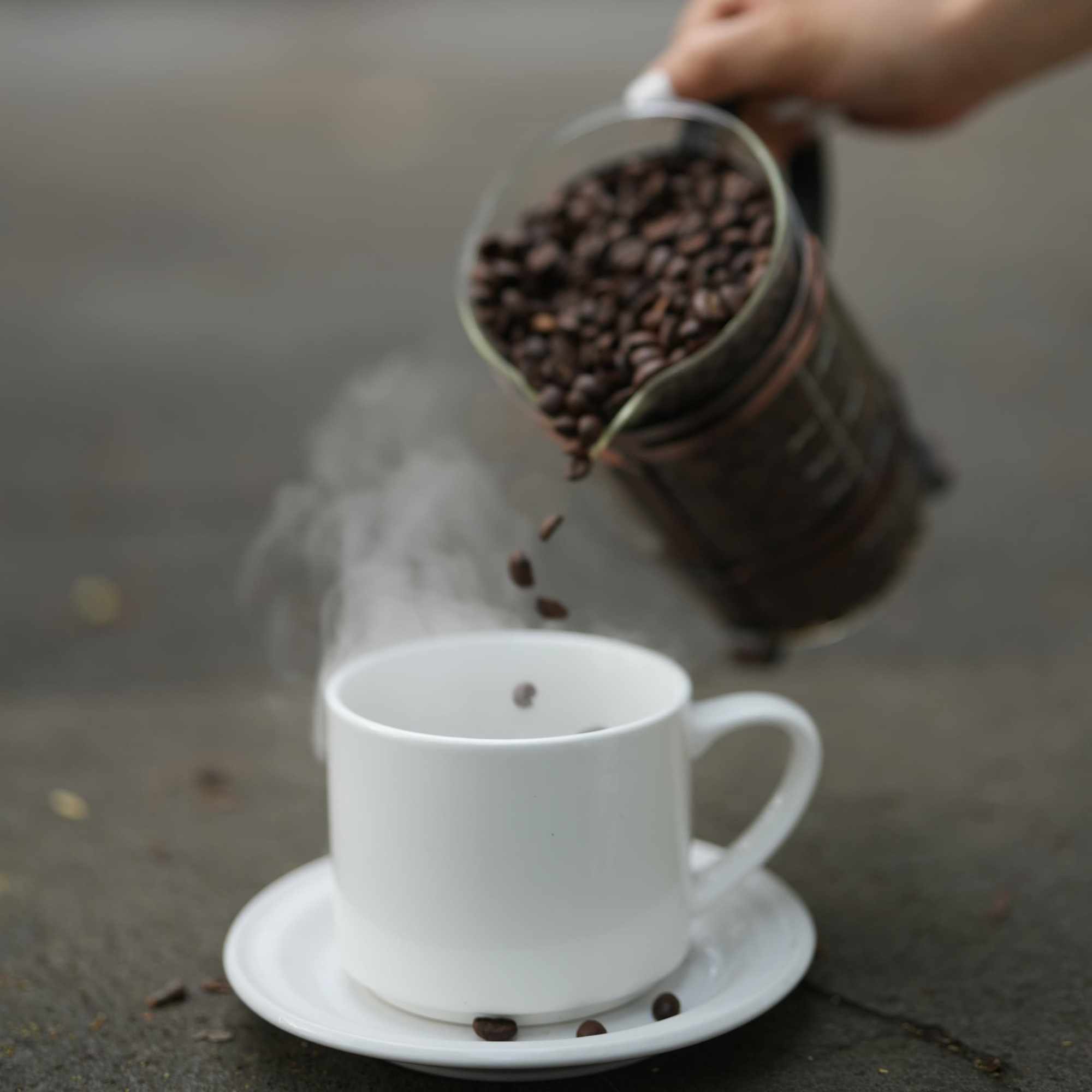 Granos de café cayendo desde una jarra hacia una taza humeante